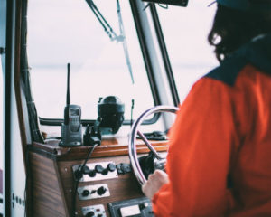 En person som står ved rattet til en mellomstor båt, sett bakfra